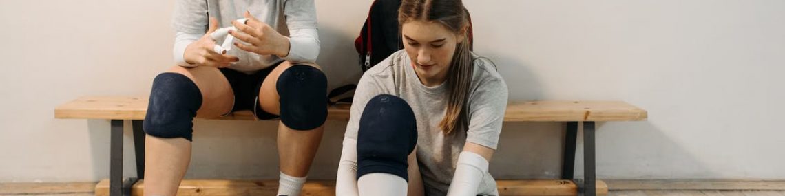 Two female volleyball players in sports gear prepare for a game, sitting on a wooden bench.